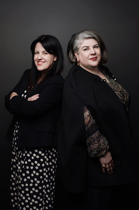 Two professionally dressed women standing back to back and smiling in front of a dark background, suggesting confidence and partnership.